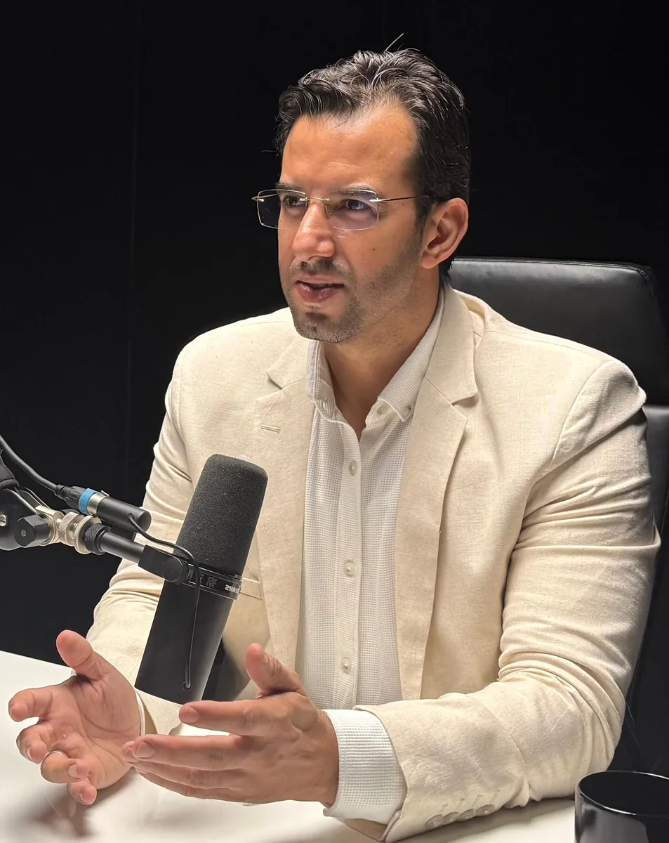 Abdelmajid Charrass en veste claire devant un microphone noir, mains ouvertes.